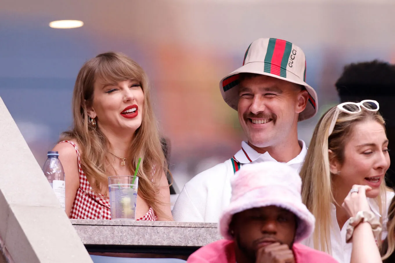 Taylor Swift and Travis Kelce smiling and sitting next to each other at the US Open in 2024. Swift is wearing a red plaid dress and Kelce is wearing a striped bucket hat and matching suspenders.