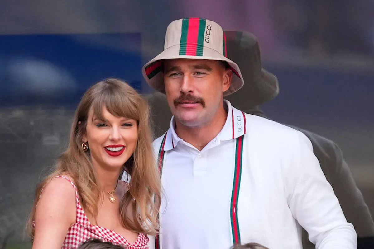 Taylor Swift and Travis Kelce at the US Open. Swift is smiling and wearing a red plaid dress. Kelce is wearing a striped bucket hat and matching suspenders over a white collared shirt.