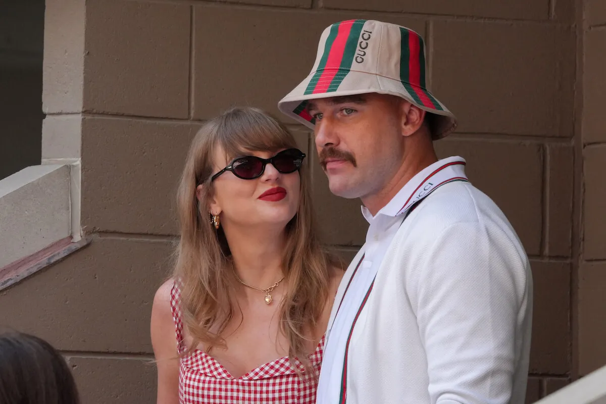 Taylor Swift and Travis Kelce at the US Open in September 2024. Swift is looking up at Kelce. She's wearing sunglasses and a red plaid dress. Kelce is wearing a plaid hat and matching suspenders over a white shirt.