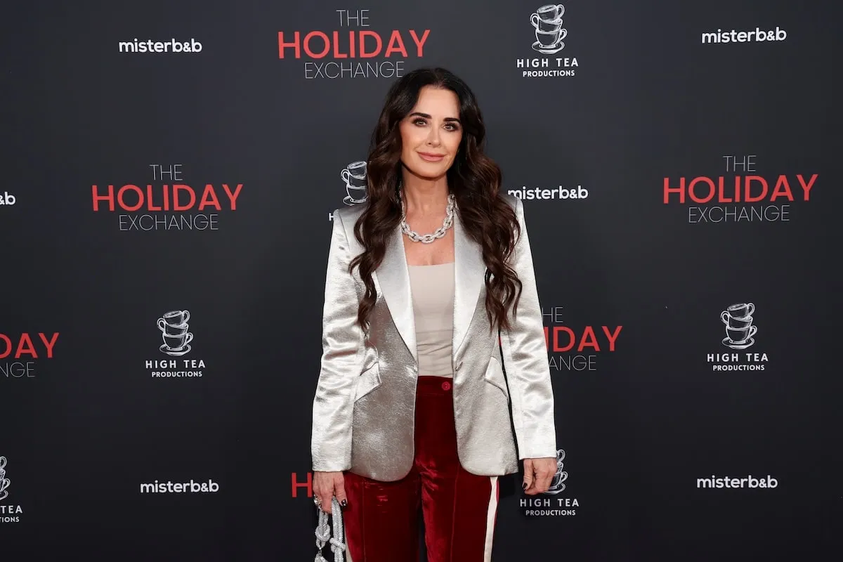 Wearing a silver blazer and red velvet pants, Kyle Richards stands on the red carpet at "The Holiday Exchange" premiere