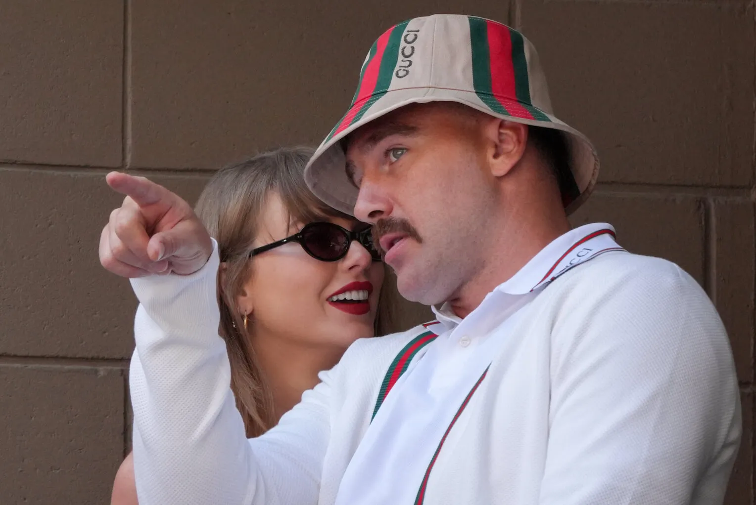 Travis Kelce and Taylor Swift at the US Open in September 2024. Kelce is pointing and looking in the distance as Swift leans in toward his ear.