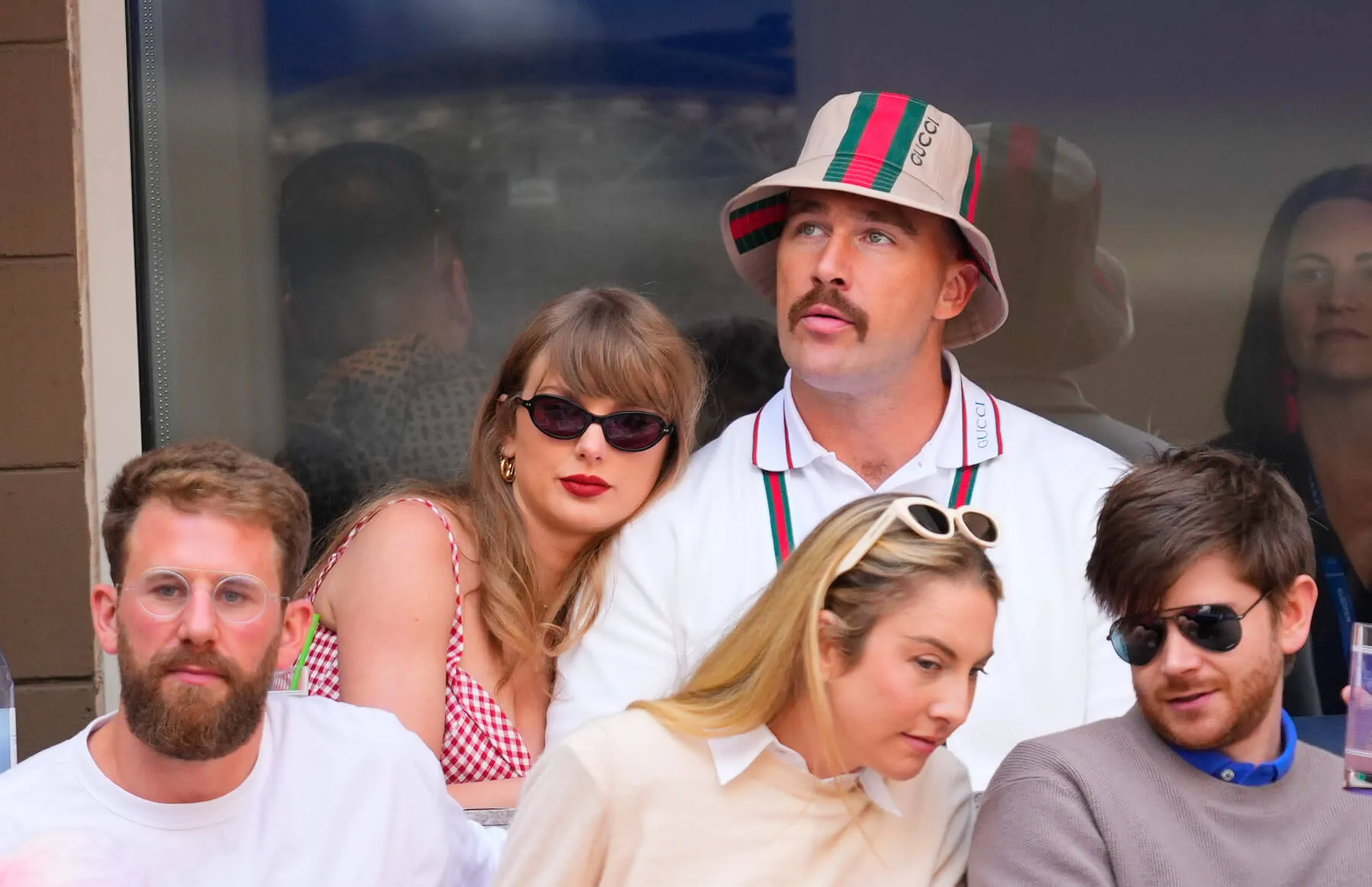 Taylor Swift leaning on Travis Kelce at the US Open. Swift is wearing a plaid dress and sunglasses and Kelce is wearing a striped bucket hat and matching suspenders.