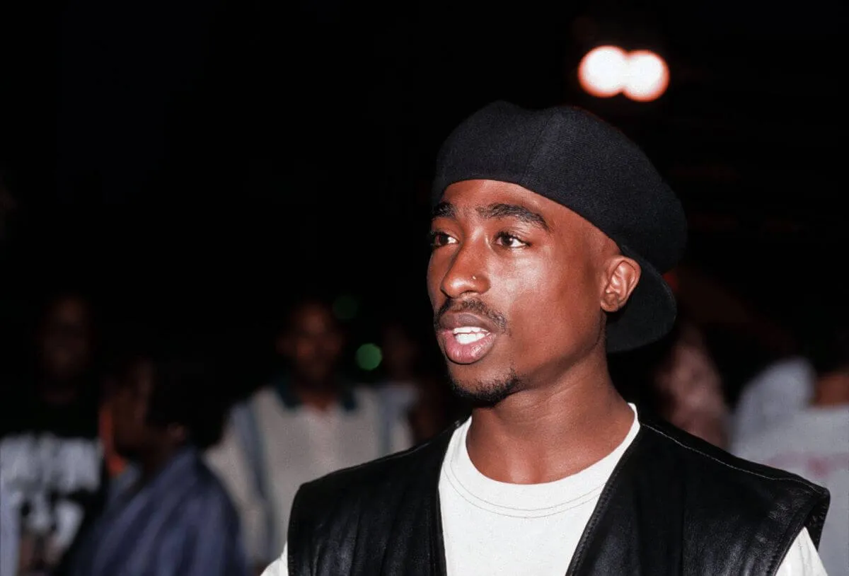 Tupac Shakur wears a hat and a black vest over a white shirt.