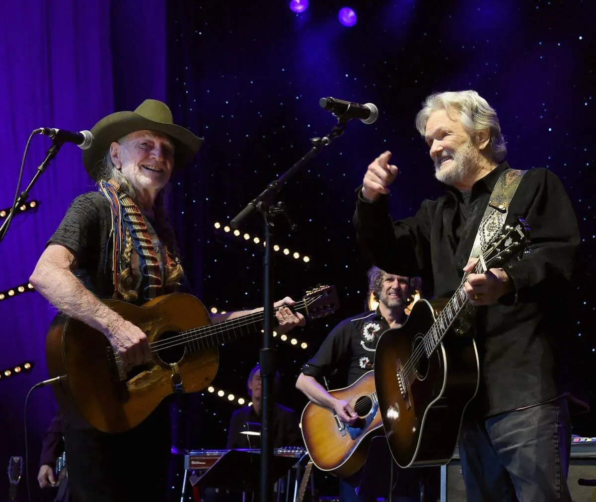 Willie Nelson and Kris Kristofferson hold guitars and stand on stage together. Nelson wears a cowboy hat.