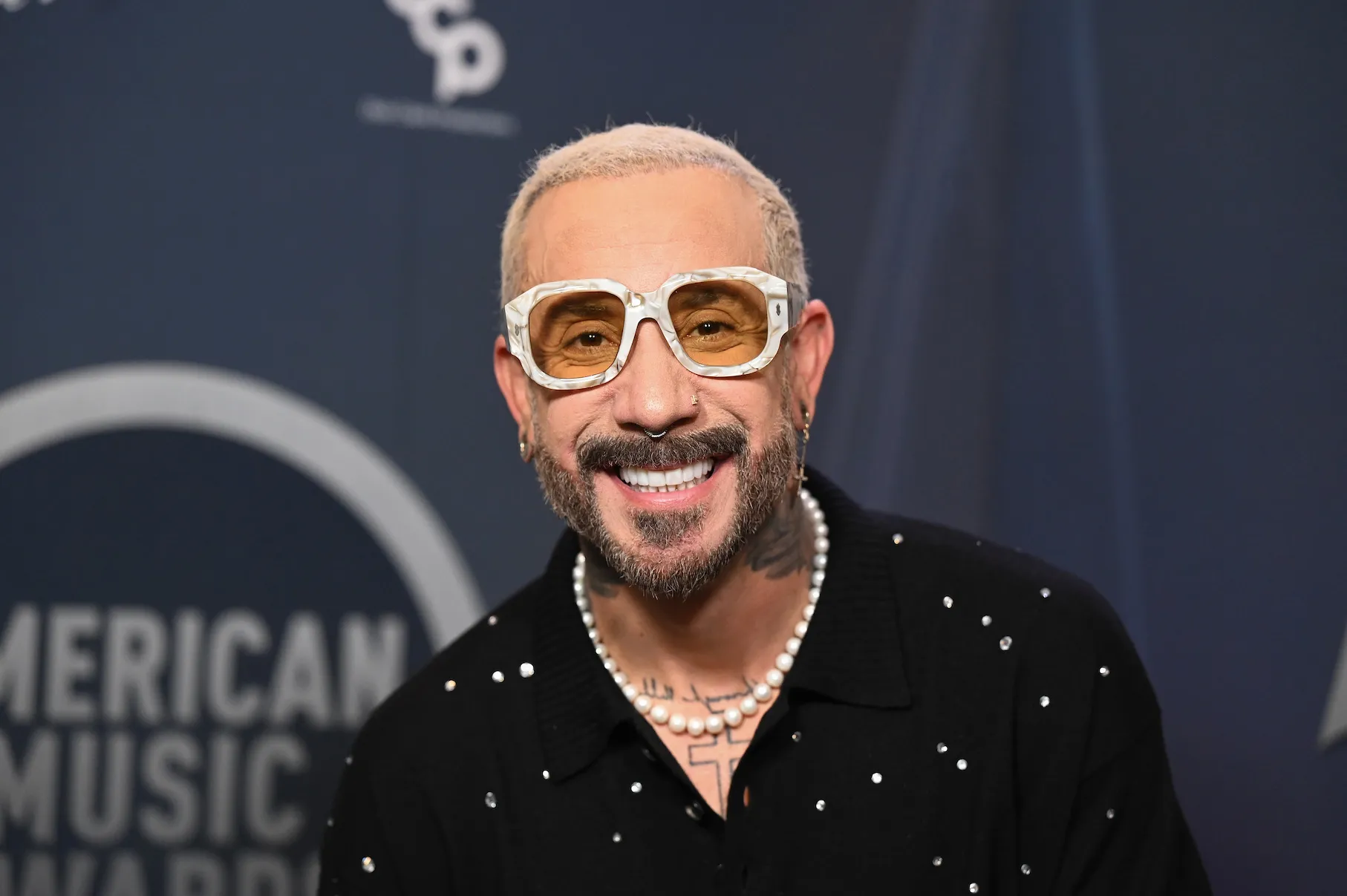 AJ McLean smiling for the camera. He's wearing white-rimmed glasses and a button-up.