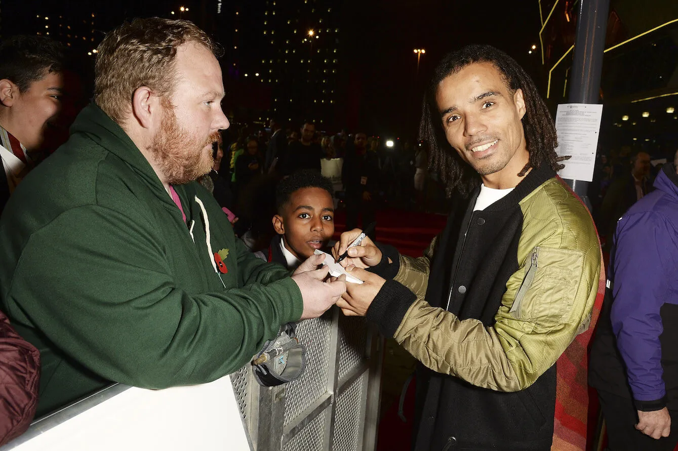 Akala signing an autograph while smiling at the camera in 2015