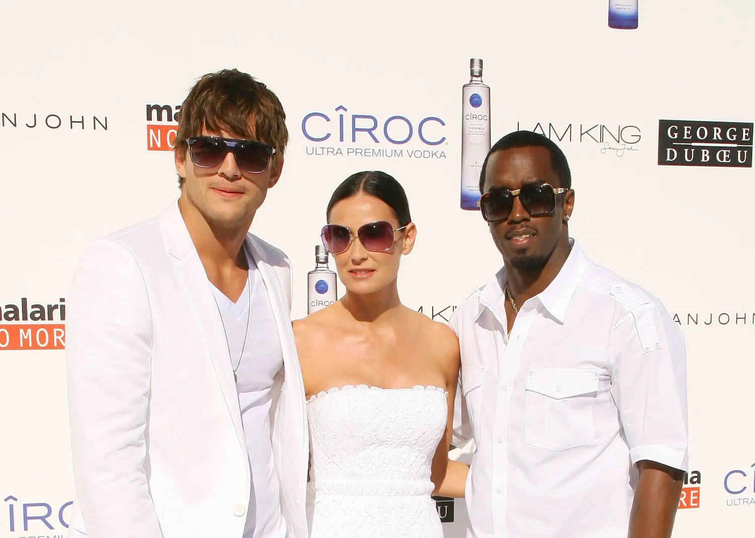 Ashton Kutcher, Demi Moore, and Sean 'Diddy' Combs at one of Combs' White Parties. They are all posing together and dressed in white with black sunglasses.