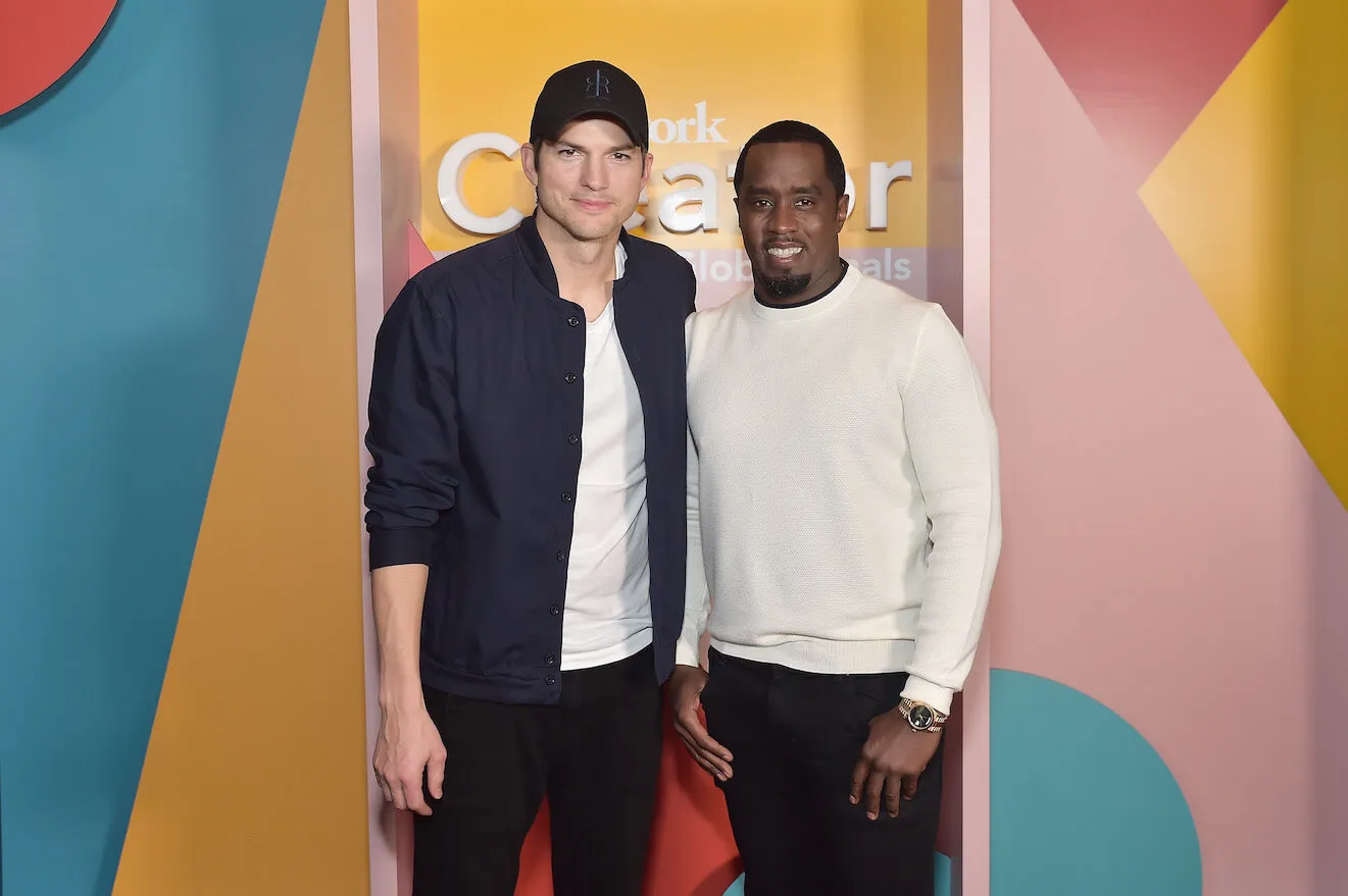 Ashton Kutcher and Sean 'Diddy' Combs standing next to each other with their arms around each other's backs. They're smiling and looking at the camera against a yellow, pink, and blue background.