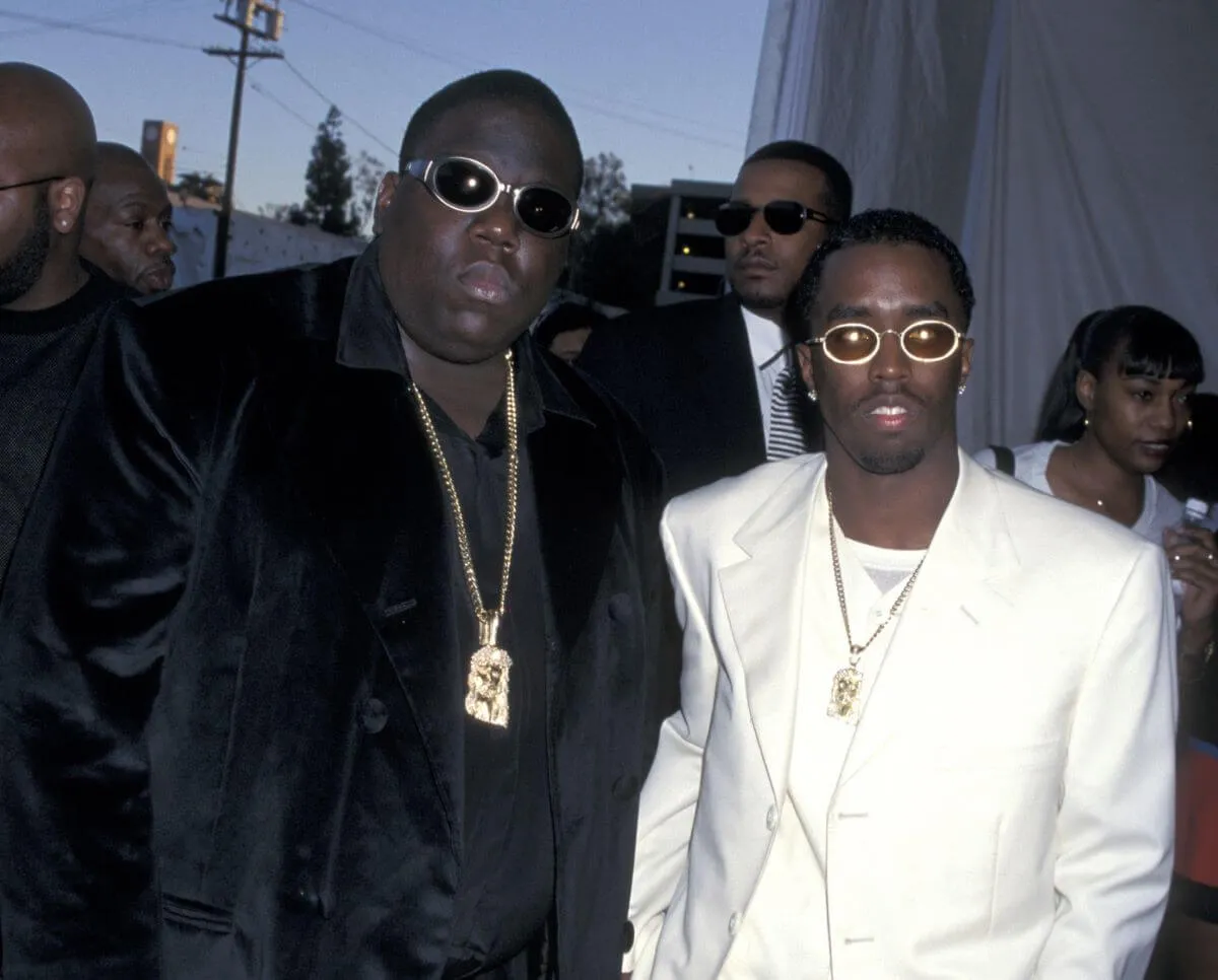 The Notorious B.I.G. and Sean 'Diddy' Combs stand together wearing sunglasses. Biggie wears a black suit and Diddy wears white.