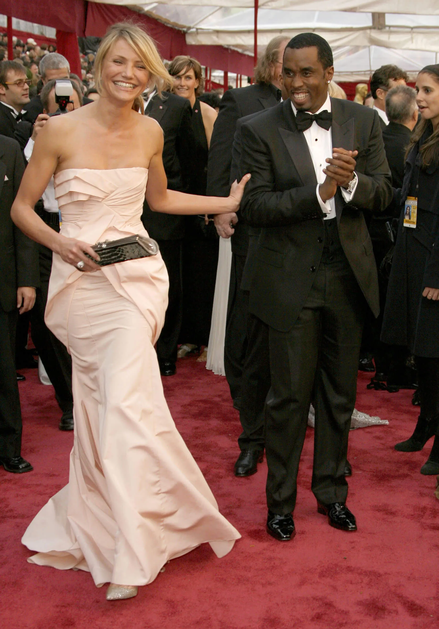 Cameron Diaz and Sean 'Diddy' Combs on the red carpet at the 80th Annual Academy Awards. Diaz is smiling and holding Combs' arm while wearing a strapless pink dress. Combs is clapping his hands together and smiling while wearing a black tux.