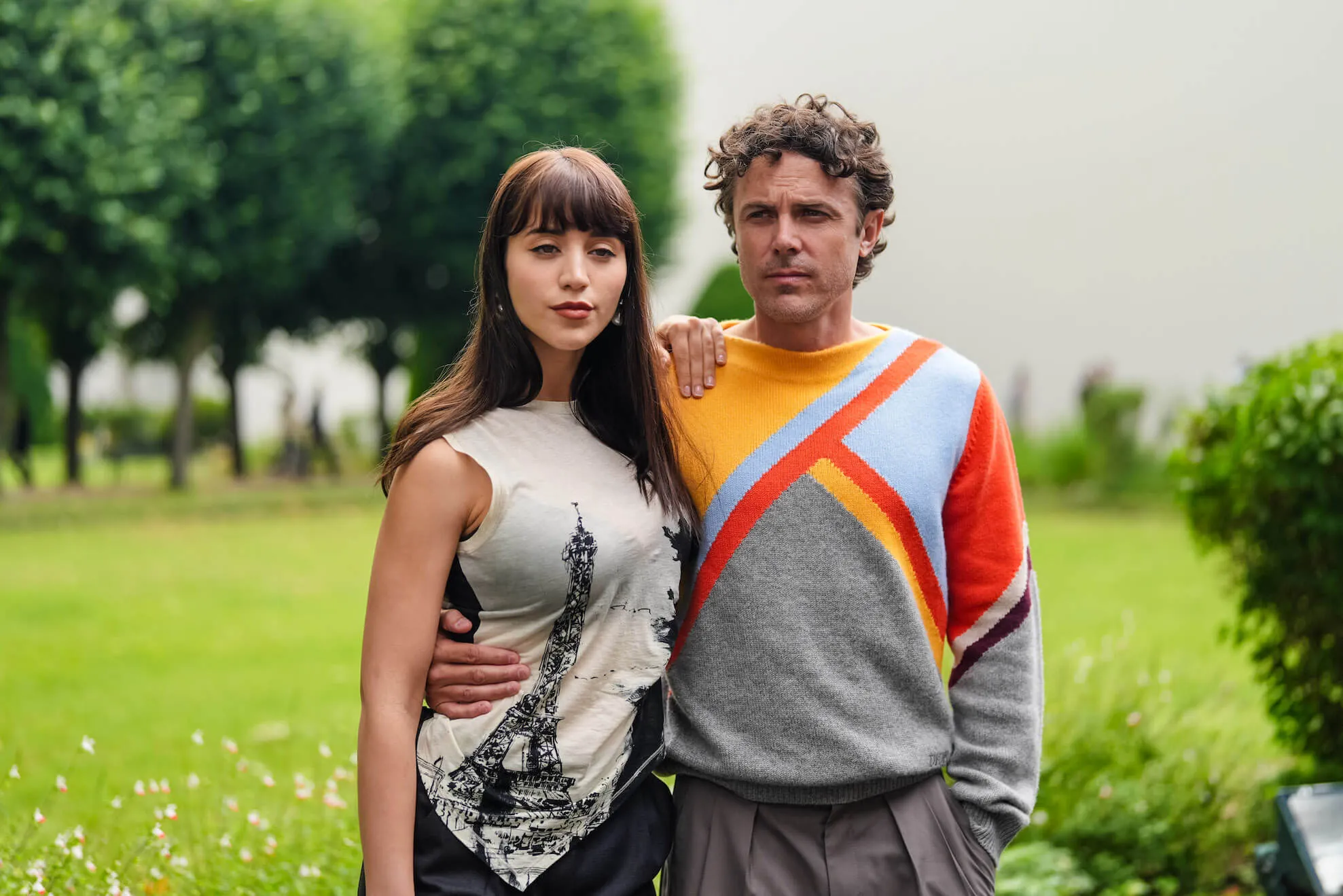 Casey Affleck wearing an orange, dark red, light blue, and gray pattern sweater with his arm around his girlfriend, Caylee Cowan, who wears a black and white tank top with Eiffel Tower print. They are posing outside.