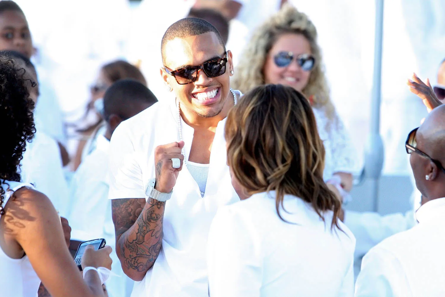 Chris Brown smiling at a White Party hosted by Sean 'Diddy' Combs. He's wearing white and surrounded by other partygoers wearing white.