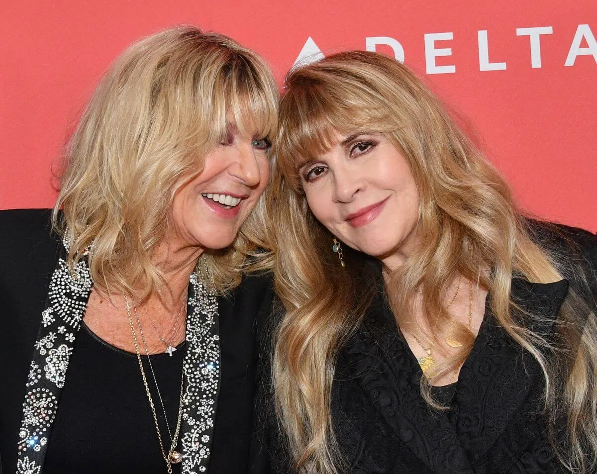 Christine McVie and Stevie Nicks wear black and stand in front of an orange background.