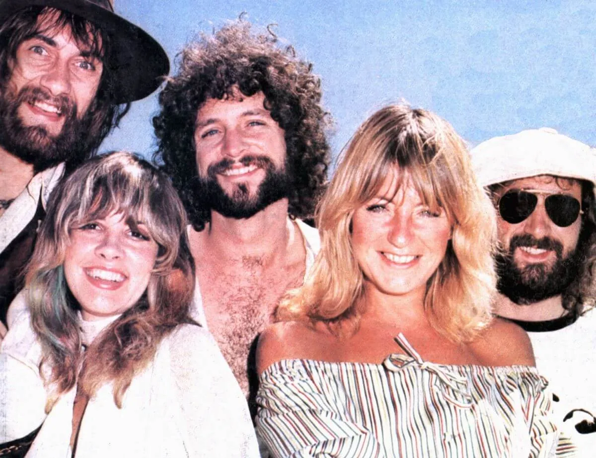 Mick Fleetwood, Stevie Nicks, Lindsey Buckingham, Christine McVie, and John McVie pose together in front of a blue background. They smile.