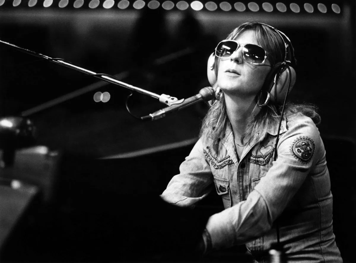 A black and white picture of Christine McVie wearing sunglasses and headphones and sitting at a piano.