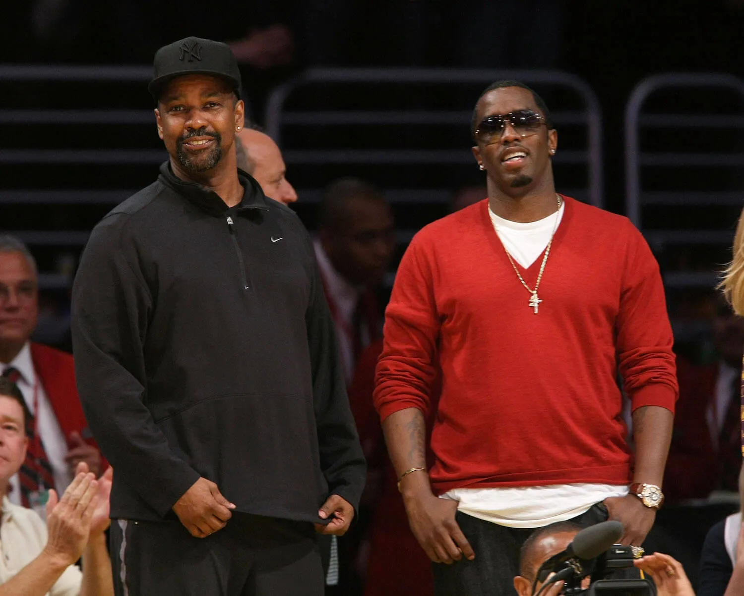 Denzel Washington wears black and stands with Sean 'Diddy' Combs at a basketball game. Diddy wears a red sweater and sunglasses.