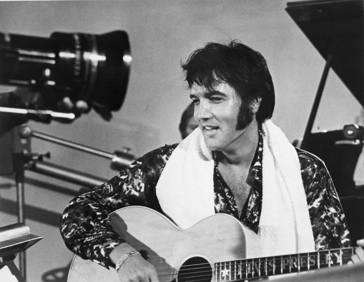 A black and white picture of Elvis with a towel around his shoulders and playing an acoustic guitar.