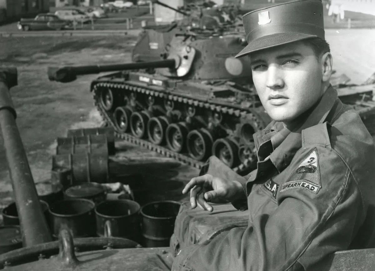 A black and white picture of Elvis Presley in his army uniform. He sits near a tank.