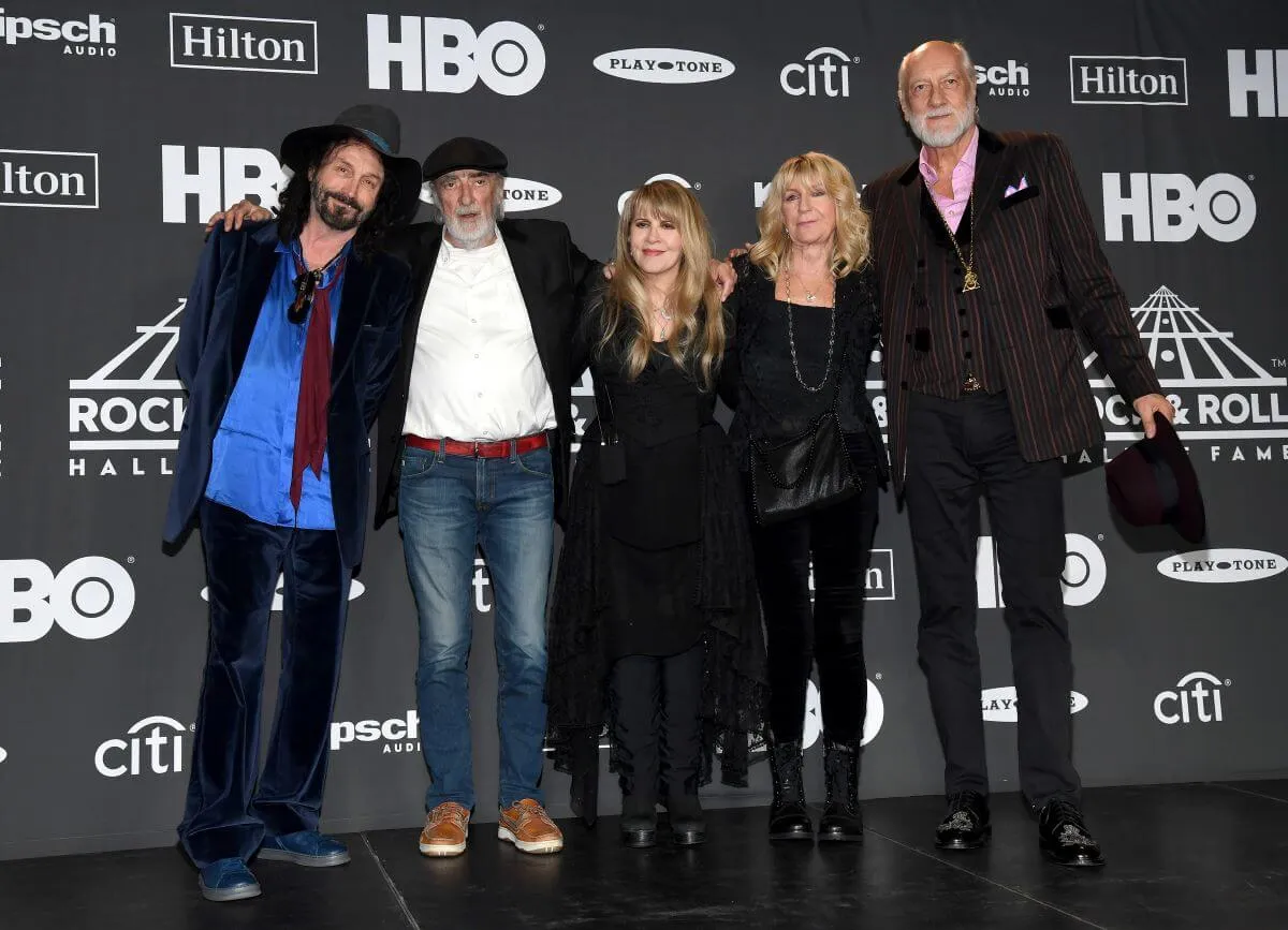 Mike Campbell, John McVie, Stevie Nicks, Christine McVie and Mick Fleetwood stand with their arms around each other.