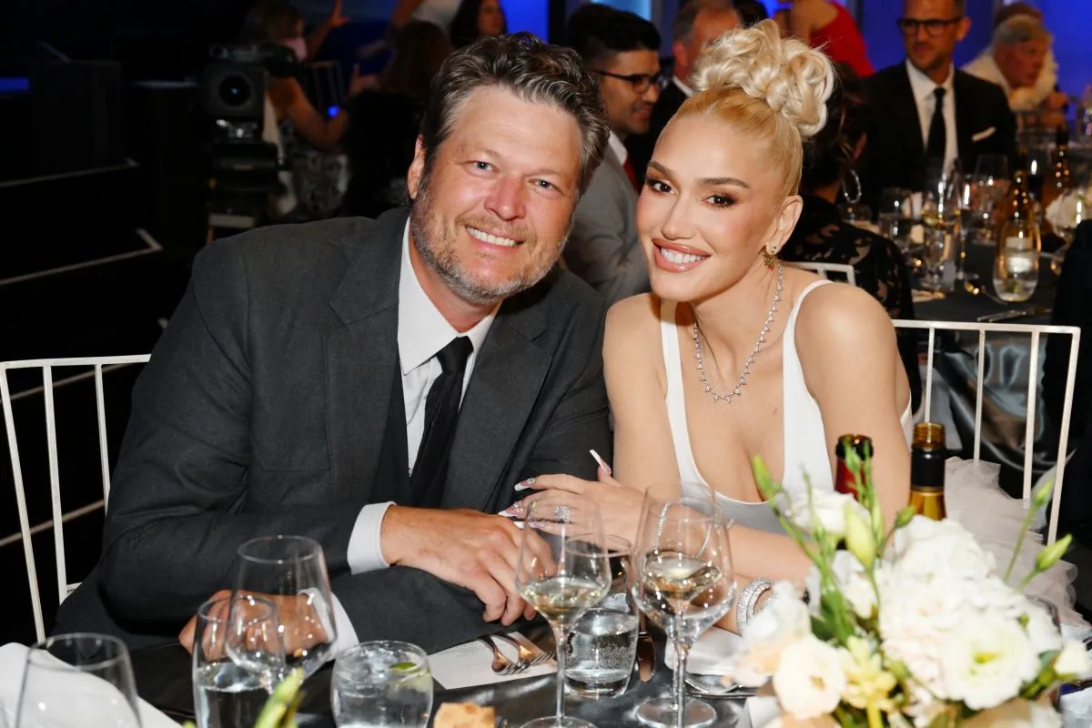 Blake Shelton and Gwen Stefani sit at a table. He wears a suit and she wears a white dress.