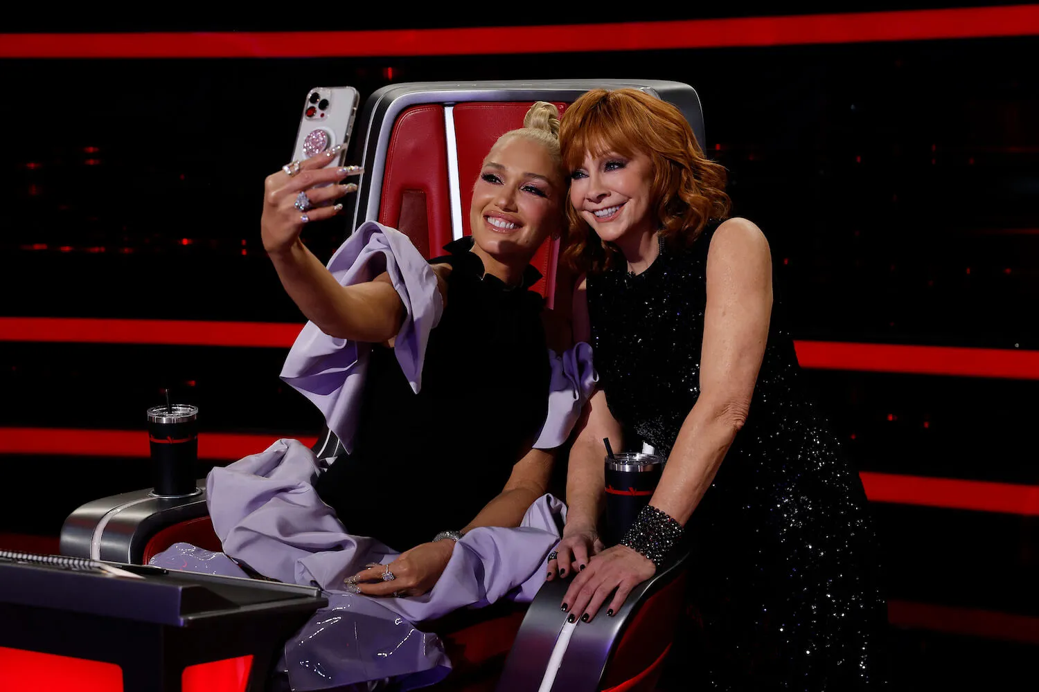 Gwen Stefani holding a phone up to take a selfie with Reba McEntire on the set of 'The Voice' Season 24. They're both smiling and posing together.