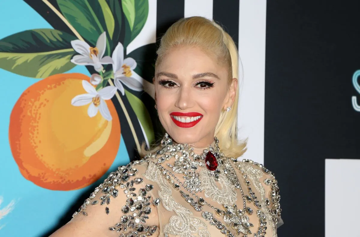 Gwen Stefani wears a rhinestoned dress and stands in front of the painting of an orange.