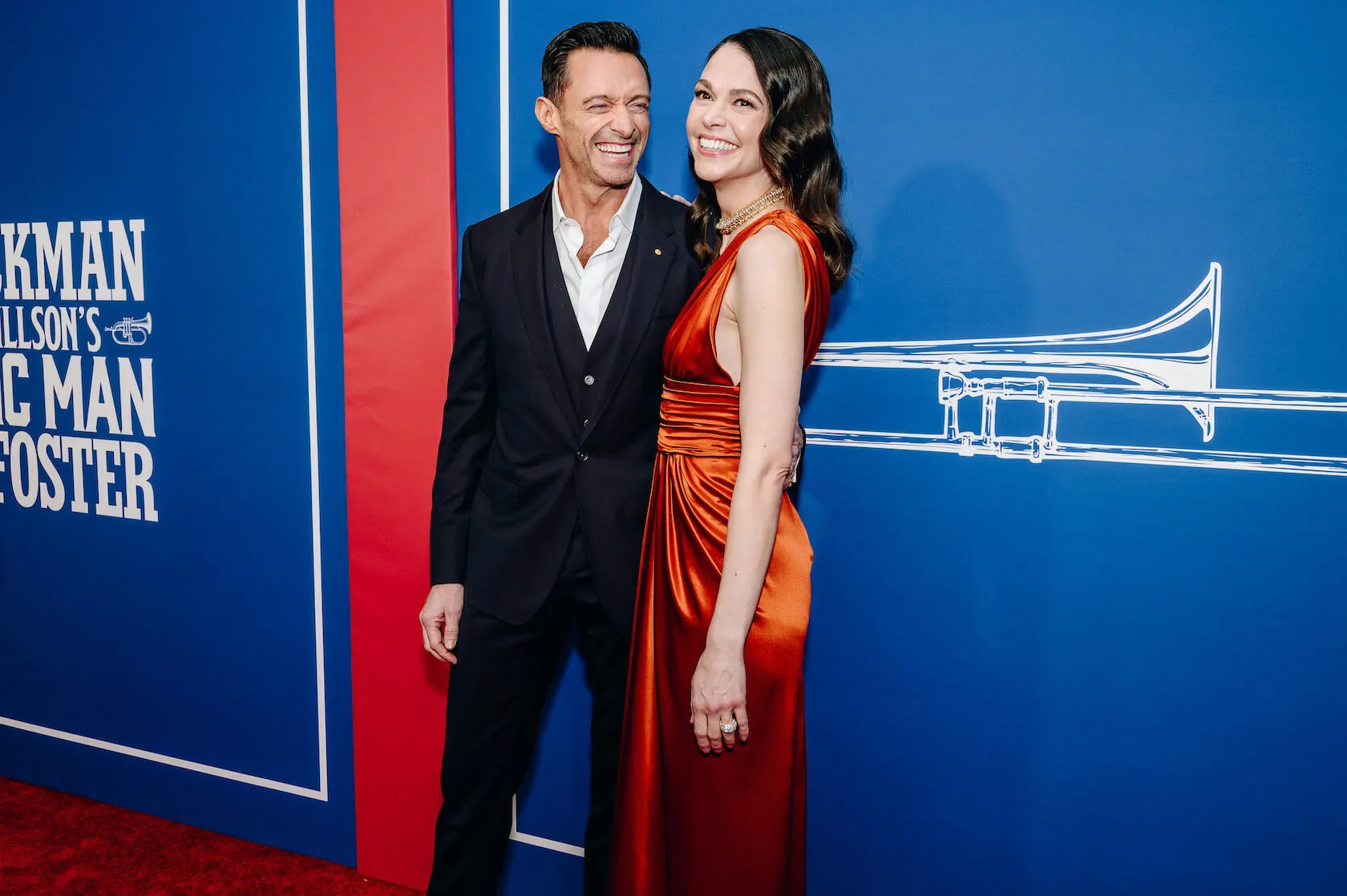 Hugh Jackman and Sutton Foster at 'The Music Man' Broadway opening in 2022. Jackman has his arm around Foster and is smiling toward her. Foster is wearing a orange dress and Jackman is wearing a suit.