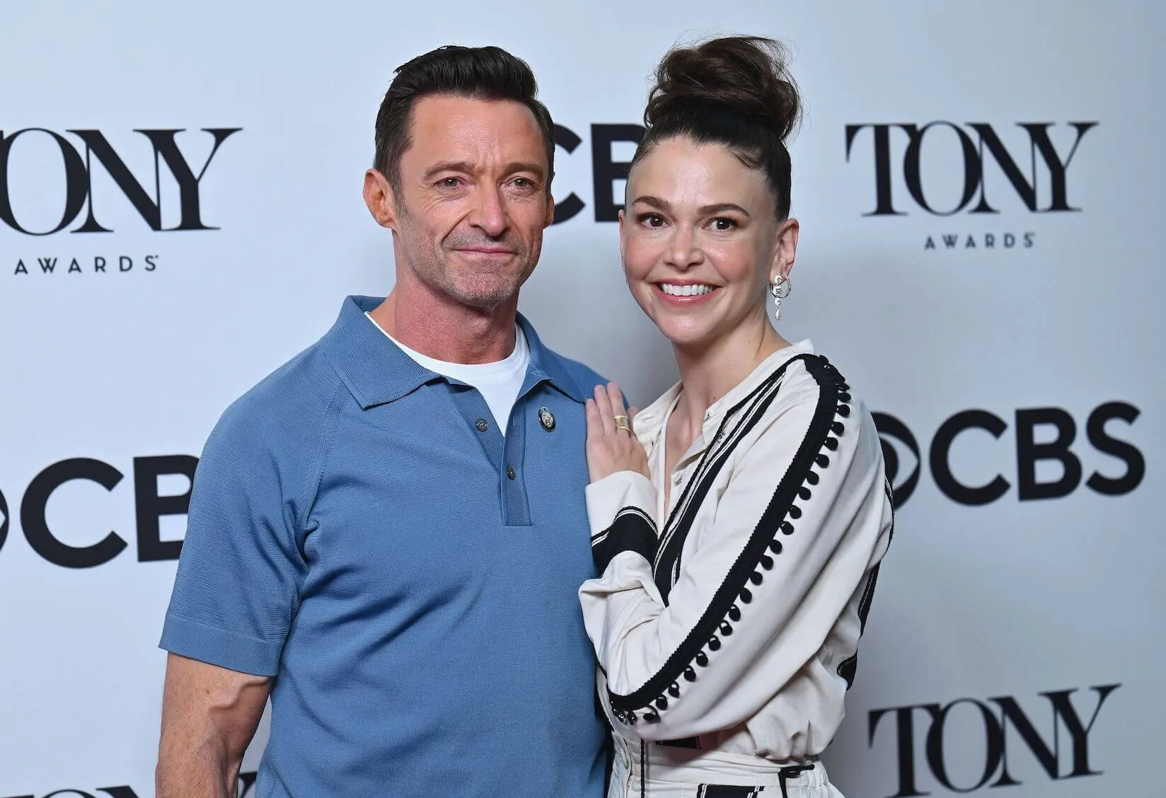 Hugh Jackman and Sutton Foster posing together at a 2022 press event. Foster has her hand against Jackman's chest as Jackman has his arm around her back.