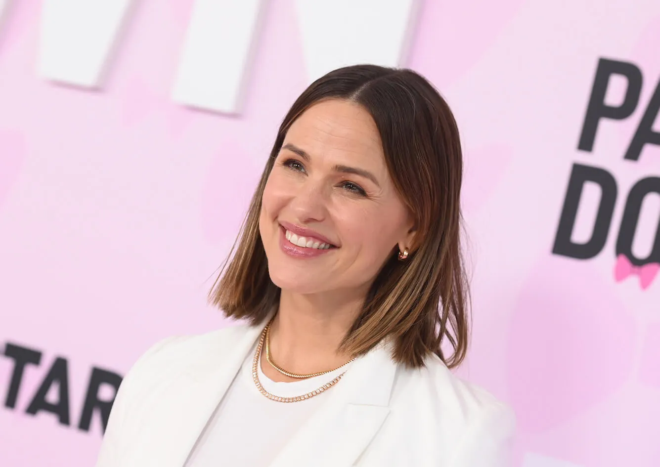 Jennifer Garner smiling at an event for 'Party Down' Season 3. She's dressed in white and standing against a pink background.