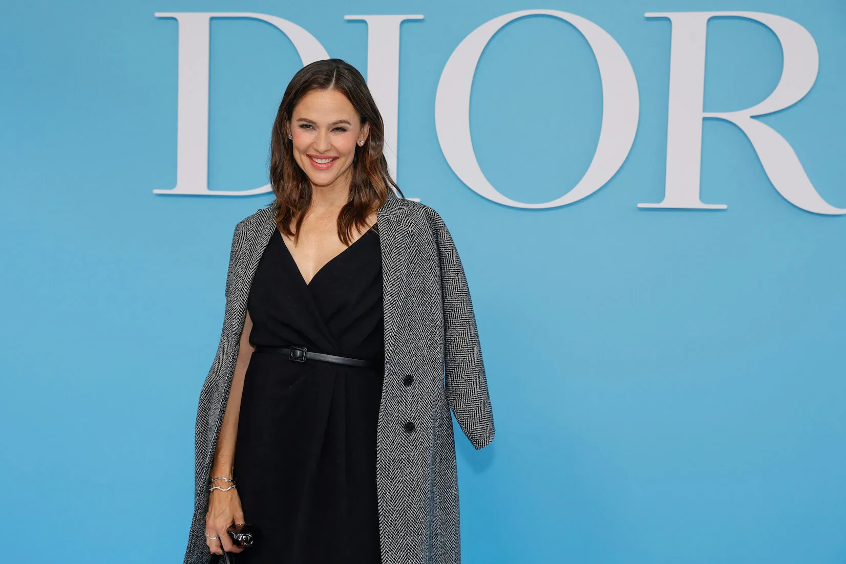 Jennifer Garner posing in a black dress and a tweed coat in front of a white Dior sign on a blue background