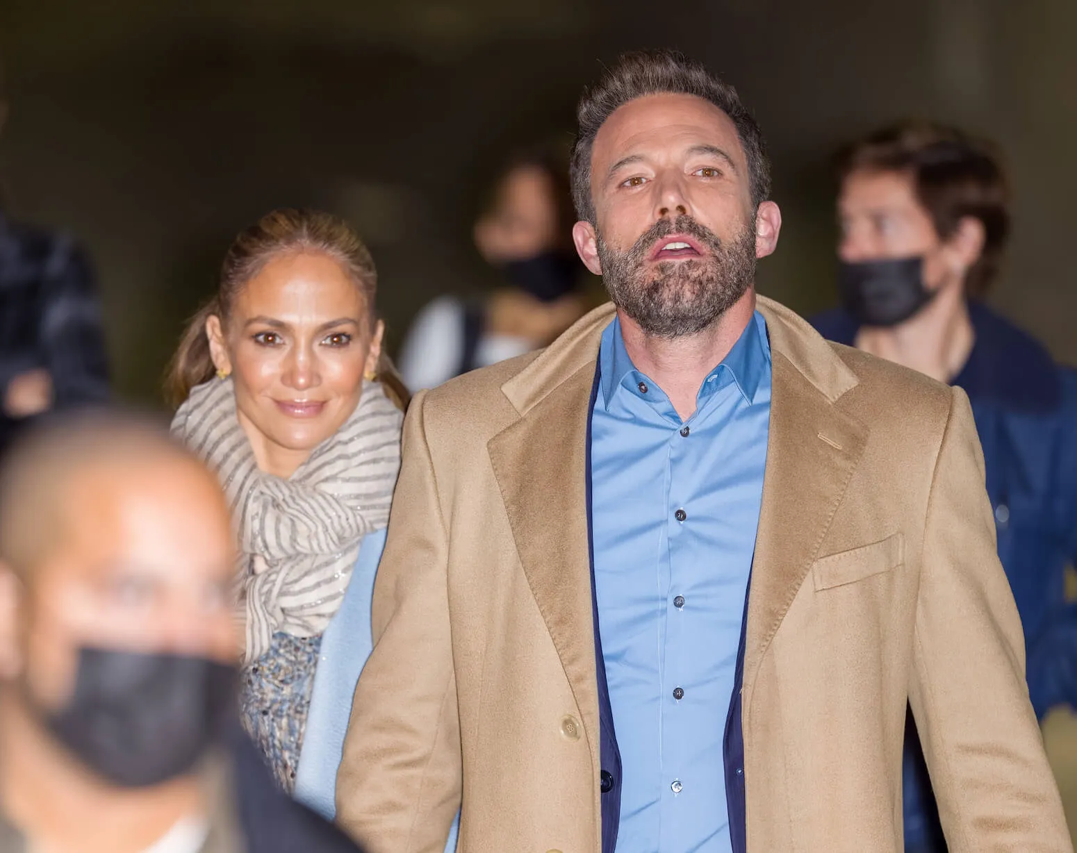 Jennifer Lopez walking in a crowd with Ben Affleck in December 2021. Lopez wears a beige scarf and Affleck wears a beige suit jacket with a blue collared shirt underneath.