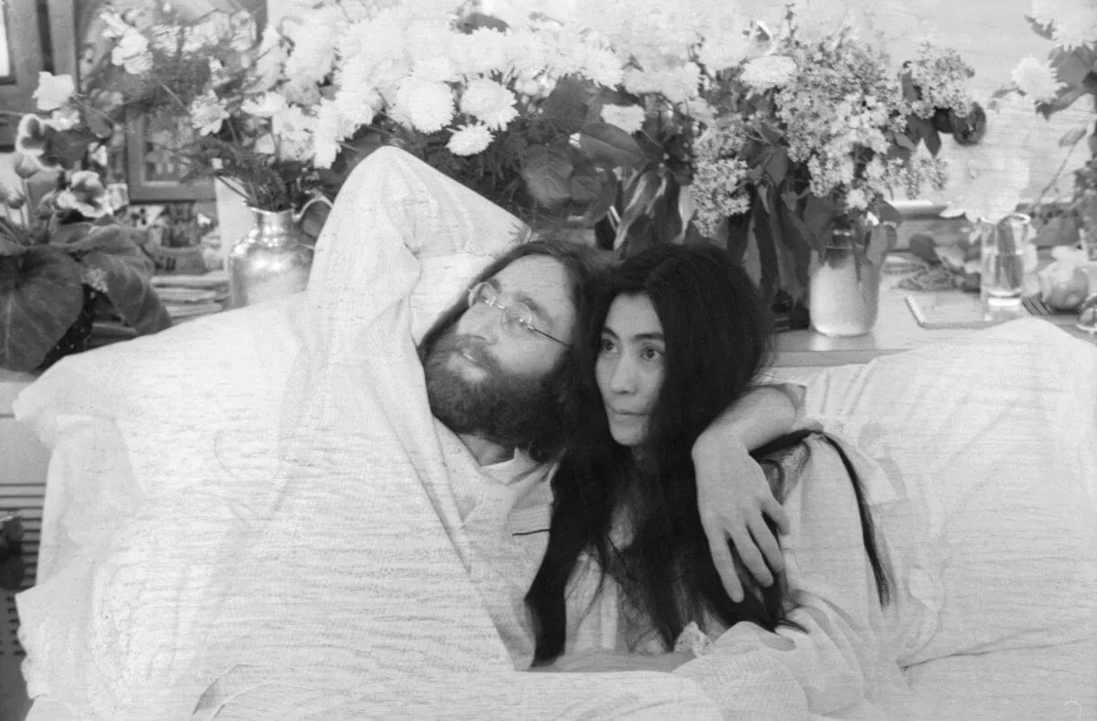 A black and white picture of John Lennon sitting in bed with his arm around Yoko Ono. There are vases of flowers behind them.