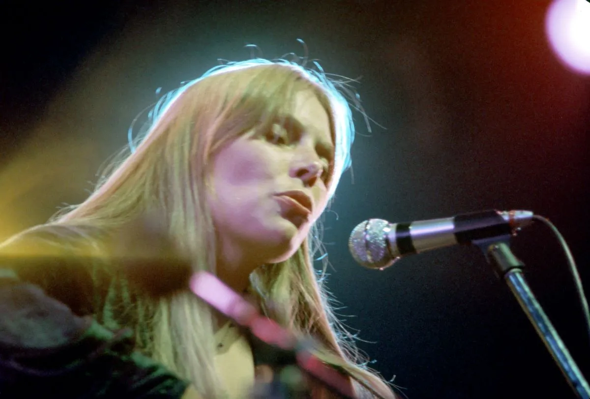 Joni Mitchell holds a guitar and sings into a microphone.