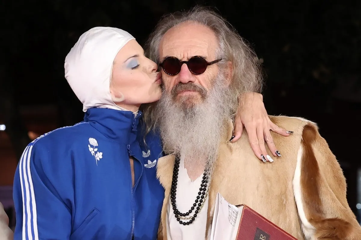 Wearing a white swim cap and blue Adidas jacket, Julia Fox kisses director Tony Kaye on the cheek at 'The Trainer' premiere