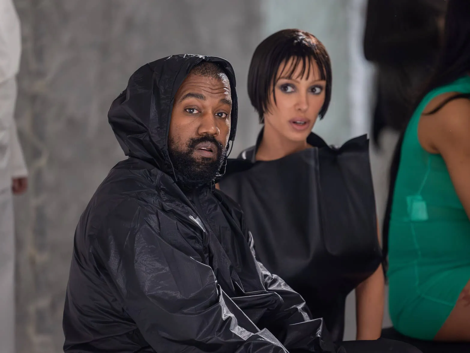 Kanye West and Bianca Censori sitting next to each other at Milan Fashion Week. West is wearing a black outfit with his hood over his head. Censori is slightly out of focus beside him in a black dress.