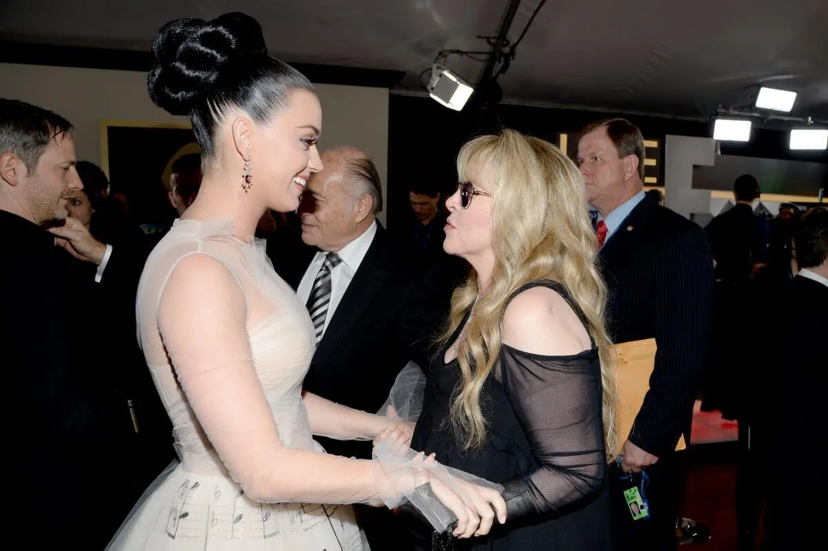 Katy Perry and Stevie Nicks hold hands and speak to each other. Perry wears a white dress and Nicks wears a black dress and sunglasses.