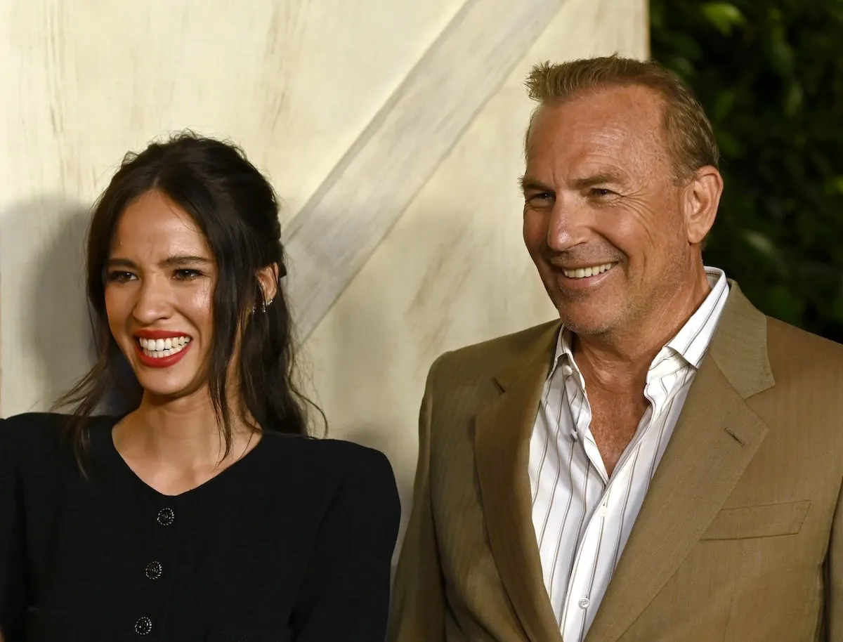 Wearing a black dress, Kelsey Asbille laughs with Kevin Costner at a media event in 2019