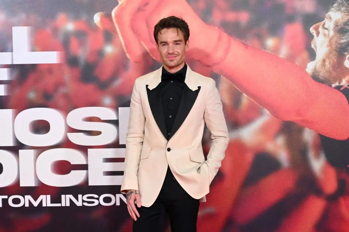 Liam Payne wears a white suit and stands in front of a poster for Louis Tomlinson's documentary.