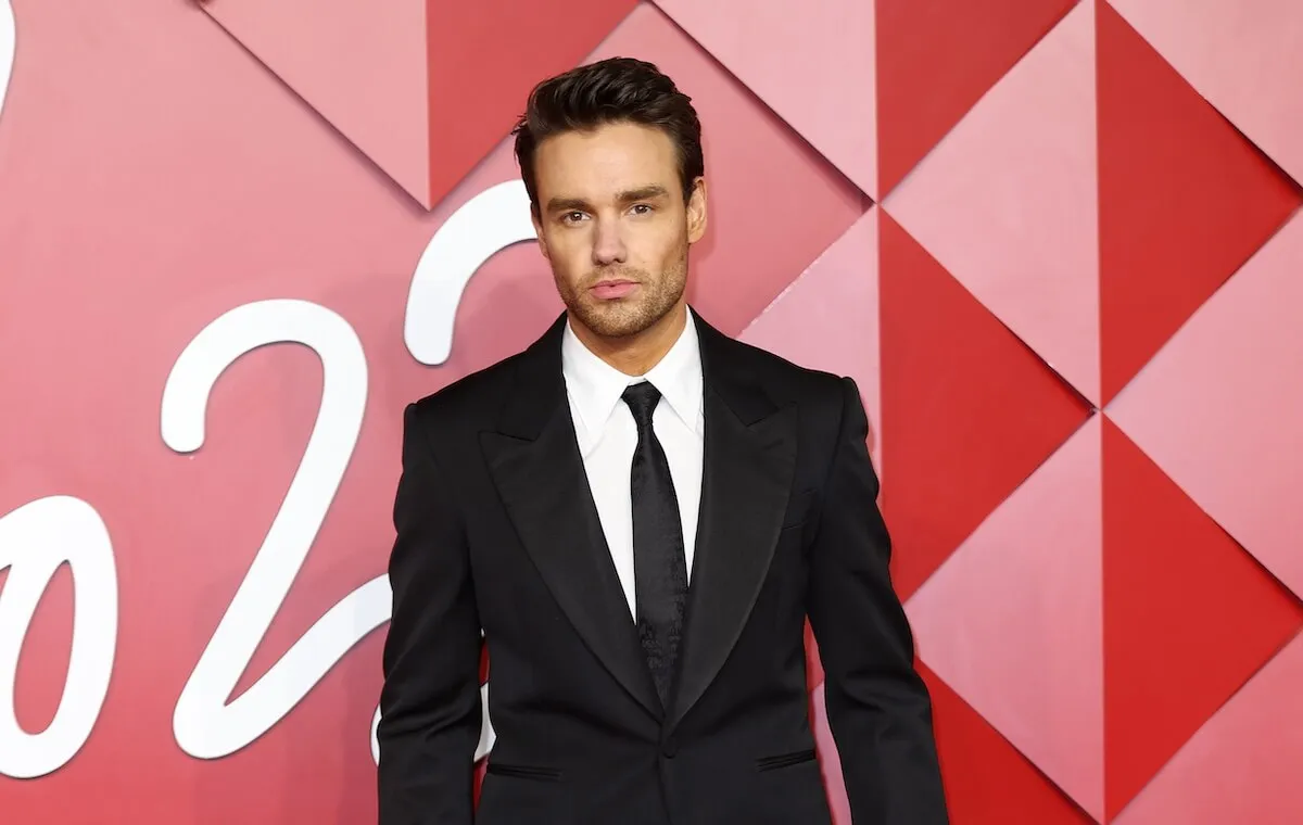 Liam Payne wears a suit and stands in front of a pink background.