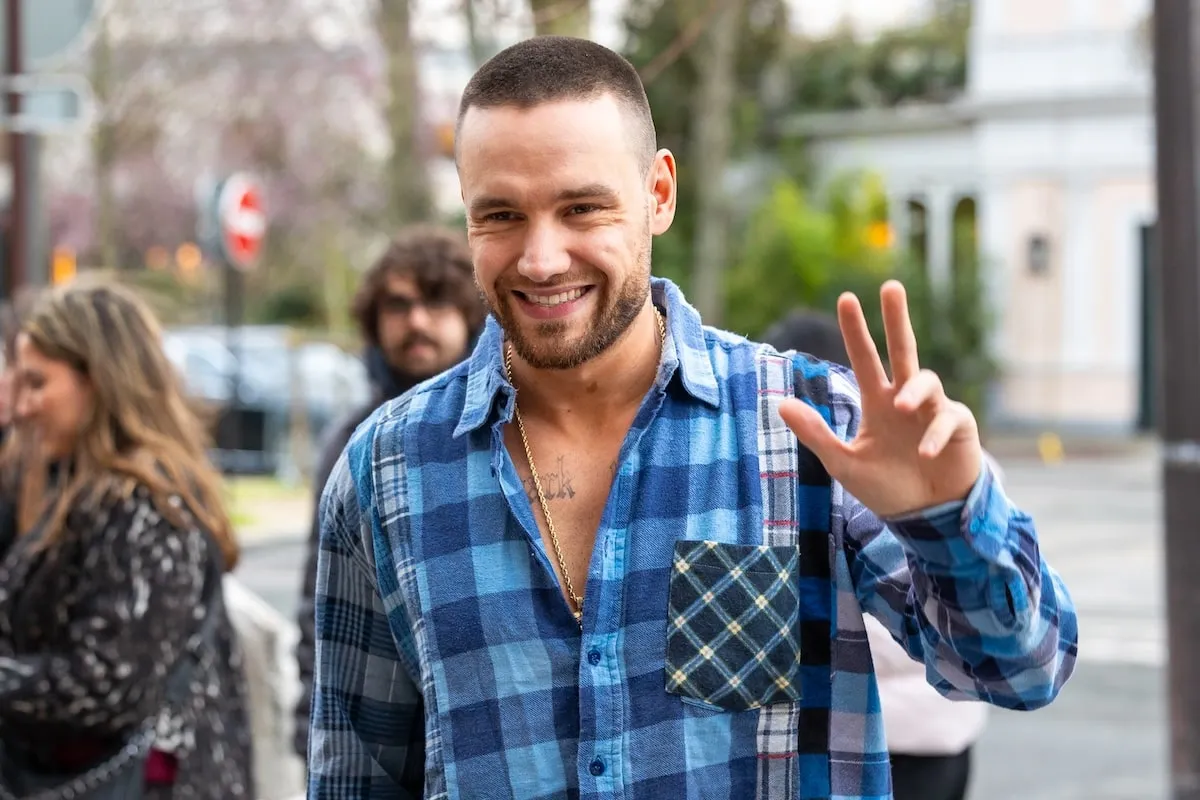 Wearing a blue plaid shirt, Liam Payne waves and smiles at fans as he walks on a Paris street