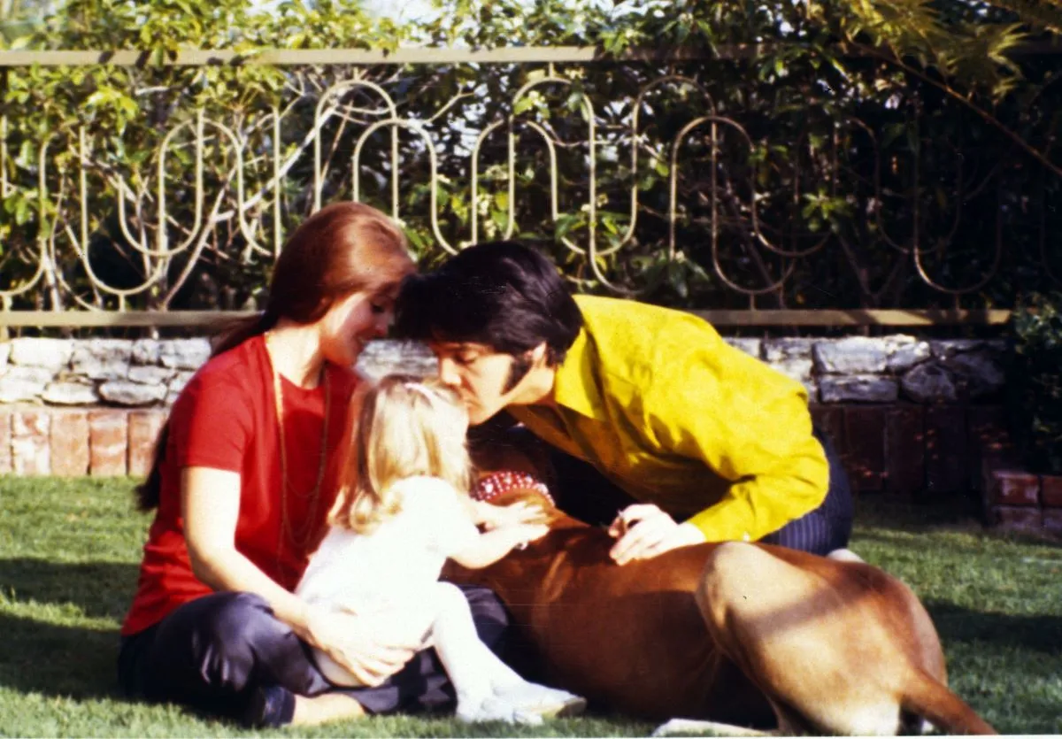 Priscilla Presley, Lisa Marie Presley, and Elvis sit outside with a dog. Elvis kisses Lisa Marie's head.