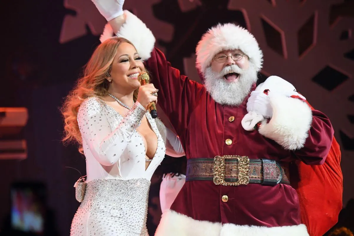 Mariah Carey wears a white dress and holds a microphone. She stands next to Santa Clause.