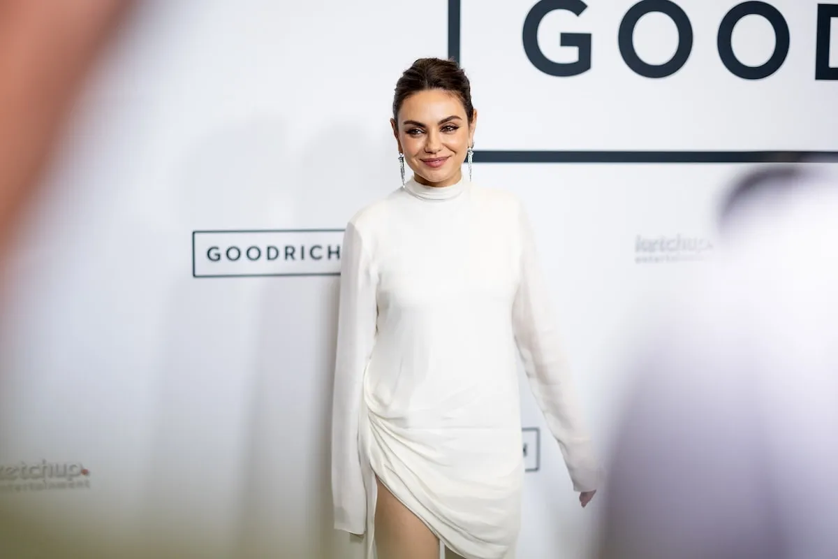 Wearing a white dress, Mila Kunis smiles as she stands alone on the red carpet