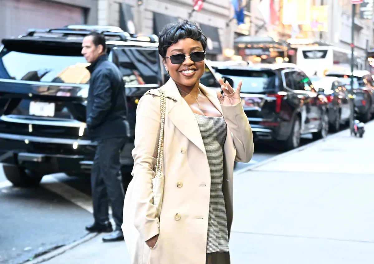 Wearing a beige trench coat and plaid mini dress, Quinta Brunson waves to cameras outside of ABC Studios