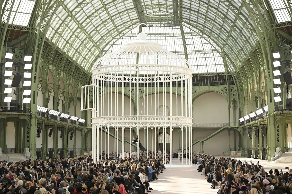 Wearing a black dress and cape, Riley Keough sits on a swing and sings during the Chanel Paris Womenswear Spring-Summer 2025 show