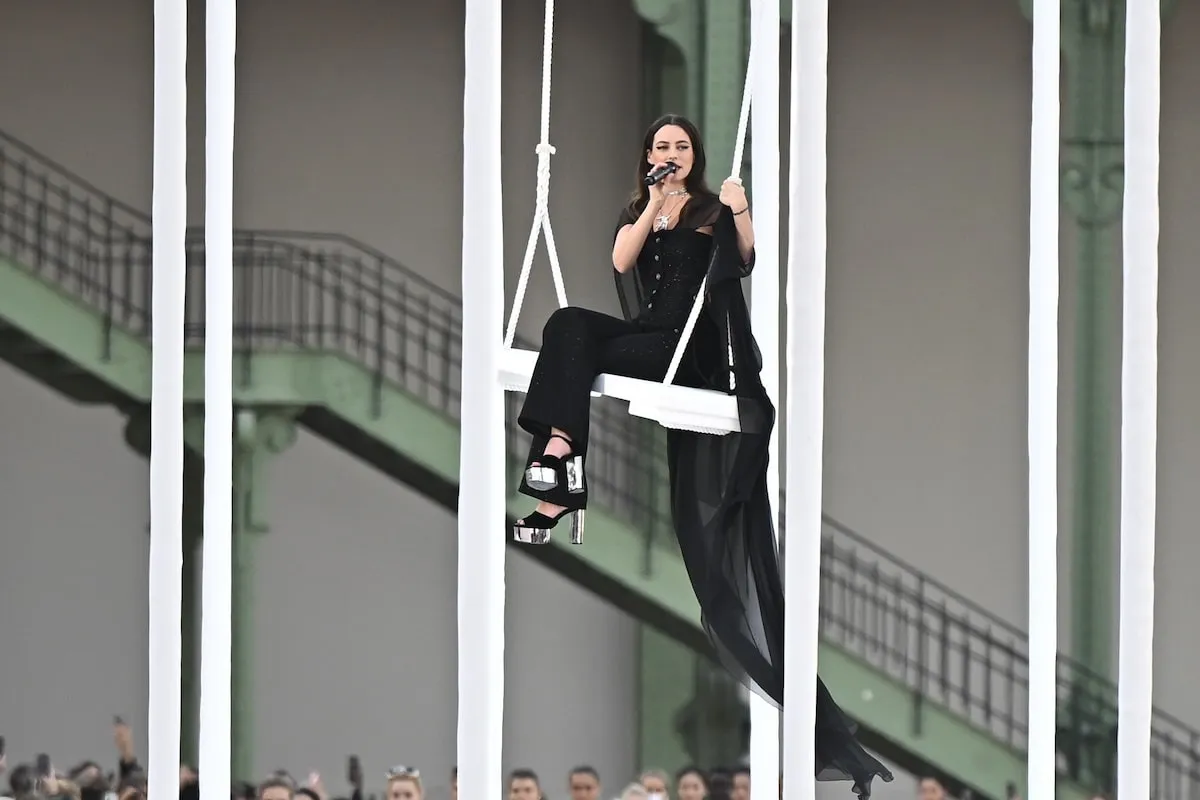 Wearing a black dress and cape, Riley Keough sits on a swing and sings during the Chanel Paris Womenswear Spring-Summer 2025 show