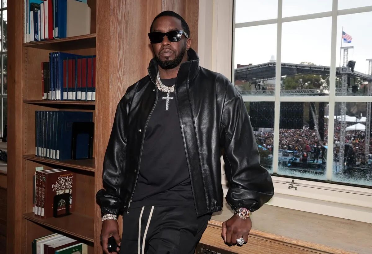 Sean 'Diddy' Combs attends event at Howard University wearing a black leather jacket and sunglasses. He stands next to a bookshelf and in front of a window.