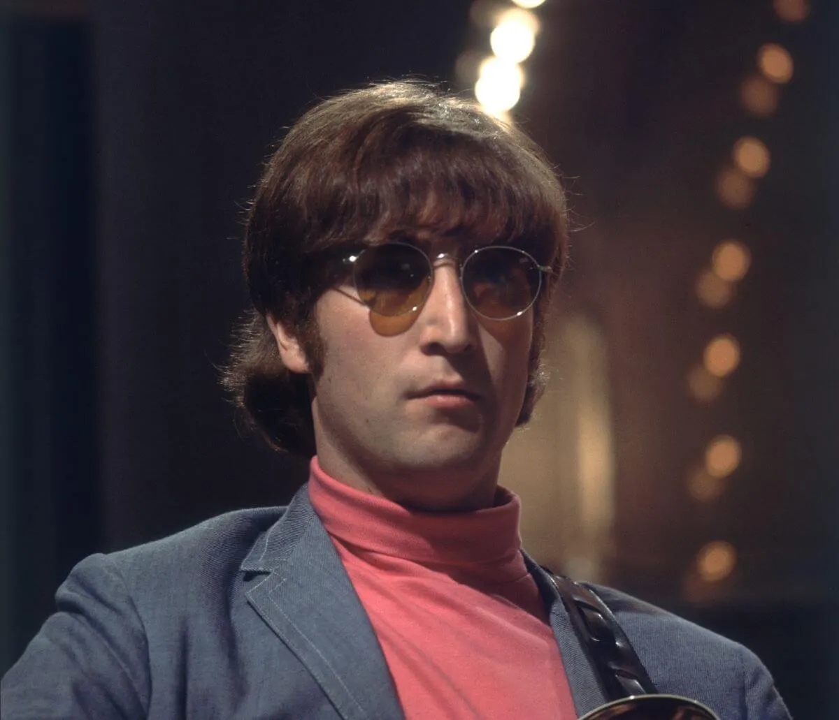 John Lennon wears a red turtleneck and a blue jacket. He wears sunglasses.