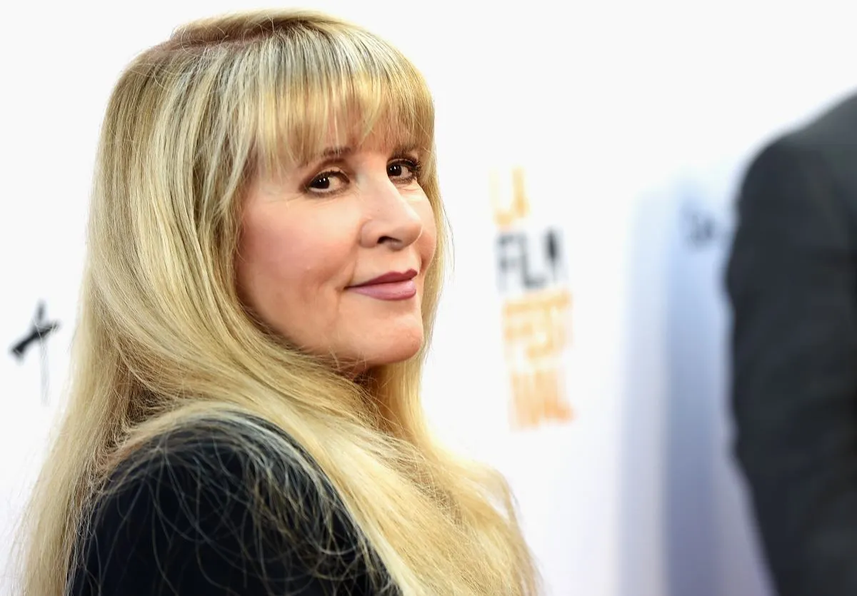 Stevie Nicks wears a black dress and stands in front of a white background.