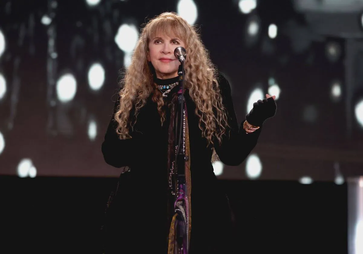 Stevie Nicks wears a black dress and stands in front of a microphone.